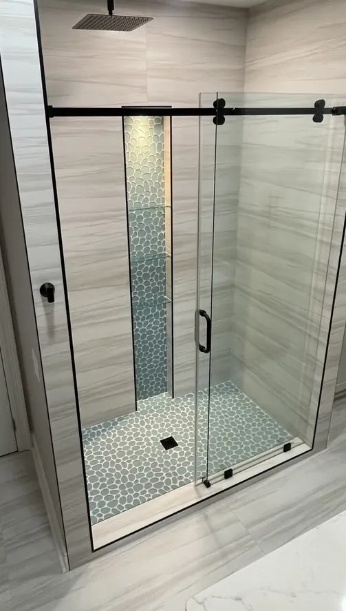 After: a modern walk-in shower with a sliding glass door, pebble-tile niche and floor, and a black rain shower